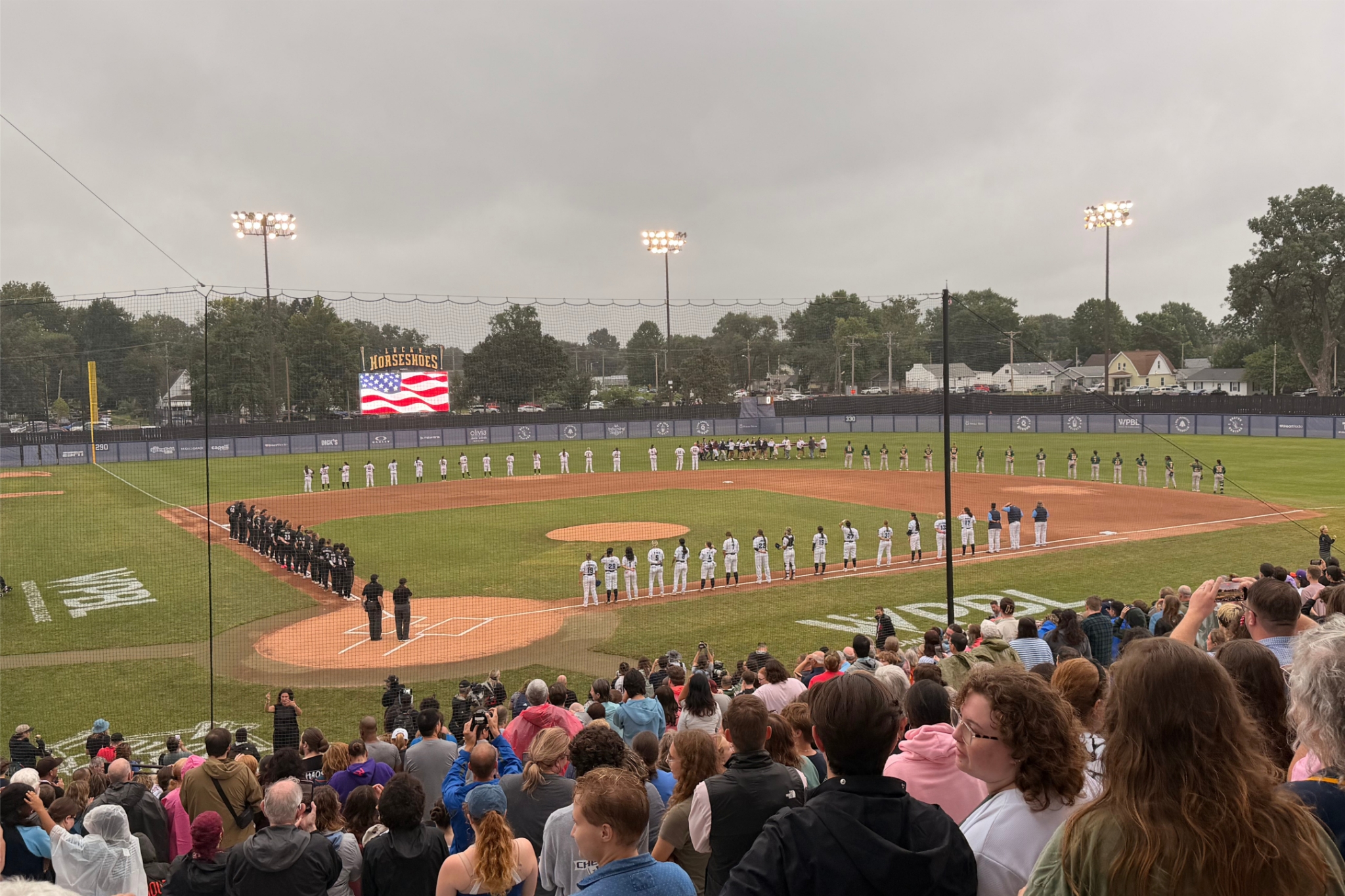Players from all four Women’s Professional Baseball League teams stand with the game’s umpires as a large American flag is unfurled before the national anthem at Robin Roberts Stadium in Springfield, Illinois.